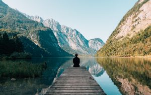 Man enjoying the view A man sitting at the pier of a lake with mountains in the near distance