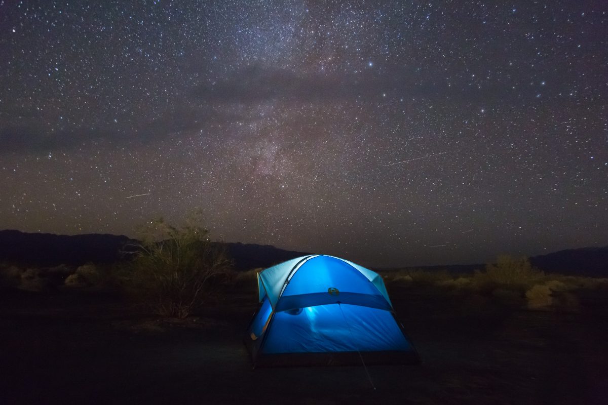 A dark blue starry night with a blue camping tent in the middle illuminated from the inside with hills and bushes in the background