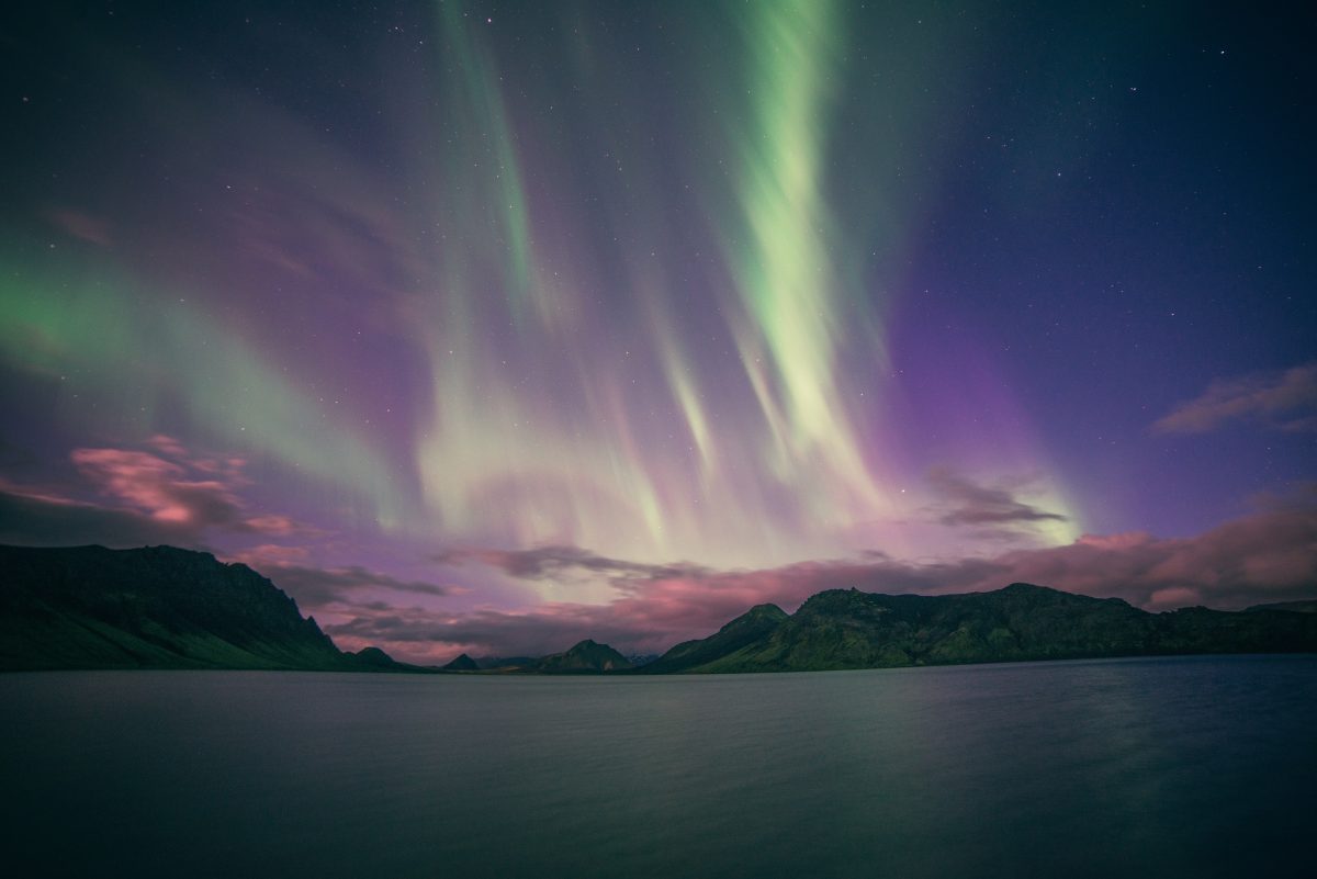 Photo of yellow green and purple aurora lights over a calm body of water with mountains and clouds in the distance in Iceland