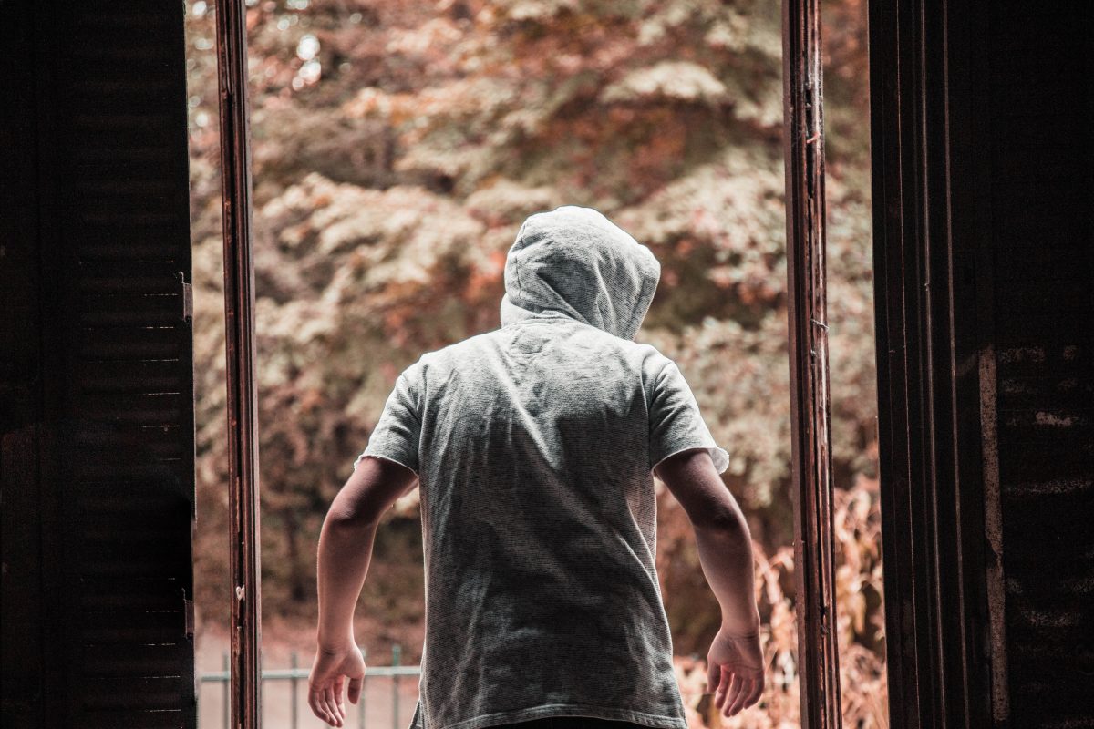 Photo of a man wearing a hooded grey shirt with his back turned towards the camera and him exiting from an entryway into sepia-colored woods