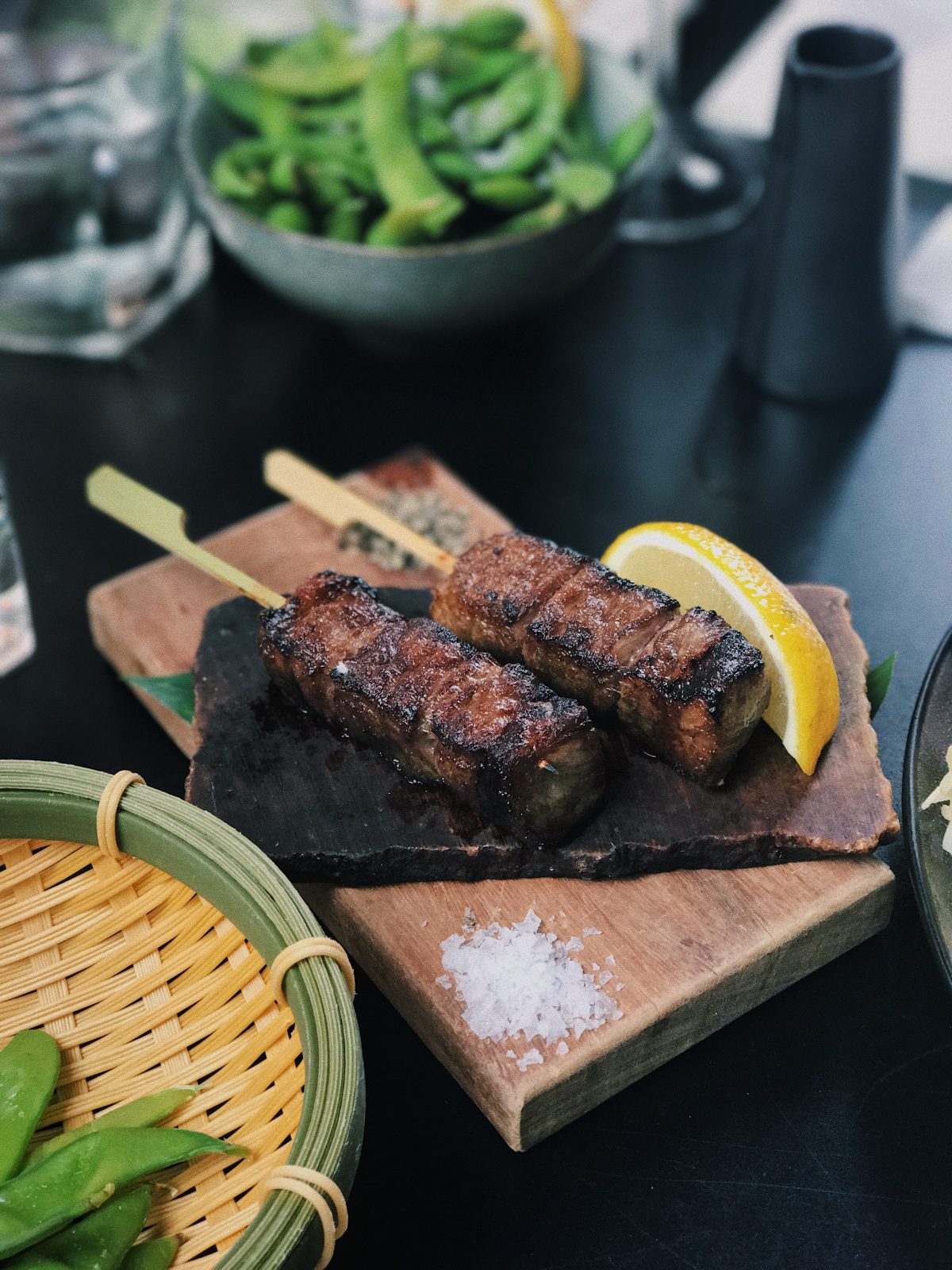 Photo of two sticks of beef chunks with three chunks per stick and a lemon slice on the side for decoration on a wooden block on top of a thicker wooden block on a black table with other bowls filled with greens on the sides with a cup of water blurred in the upper left background and a blurred dark grey container to the upper right