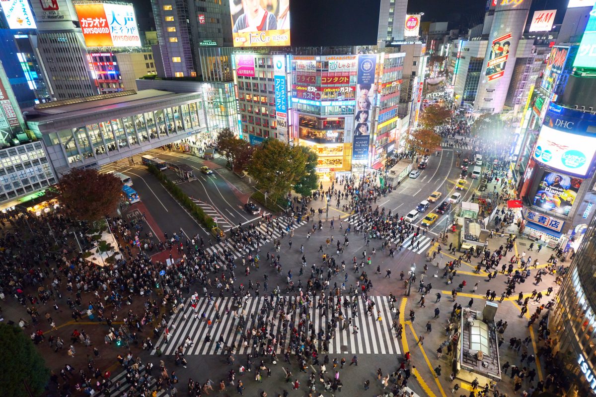 Bird’s eye view of Shibuya Crossing at night with big crowds crossing the streets and surrounding areas as lights from the buildings and signages around the area illuminate the vicinity