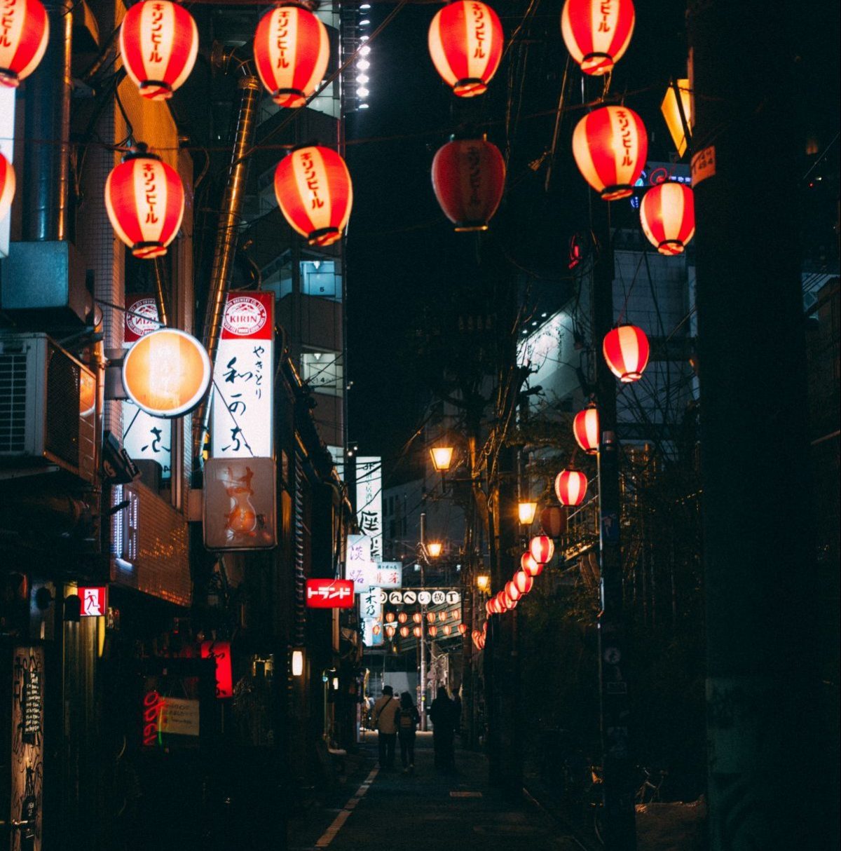 A dimly lit, small Japanese alleyway at night with red lanterns above lining the street and a few Japanese signages lit up with three people walking a few steps away