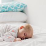 Photo of a baby with blonde hair sleeping on a bed, torso faced down, the bed has white sheets and white and blue pillows in the near distance, with the bed being against a white wall and the baby’s wearing a long-sleeved white garment with orange polka dots