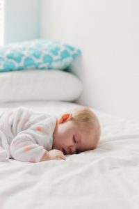 toddler sleeping Photo of a baby with blonde hair sleeping on a bed, torso faced down, the bed has white sheets and white and blue pillows in the near distance, with the bed being against a white wall and the baby’s wearing a long-sleeved white garment with orange polka dots