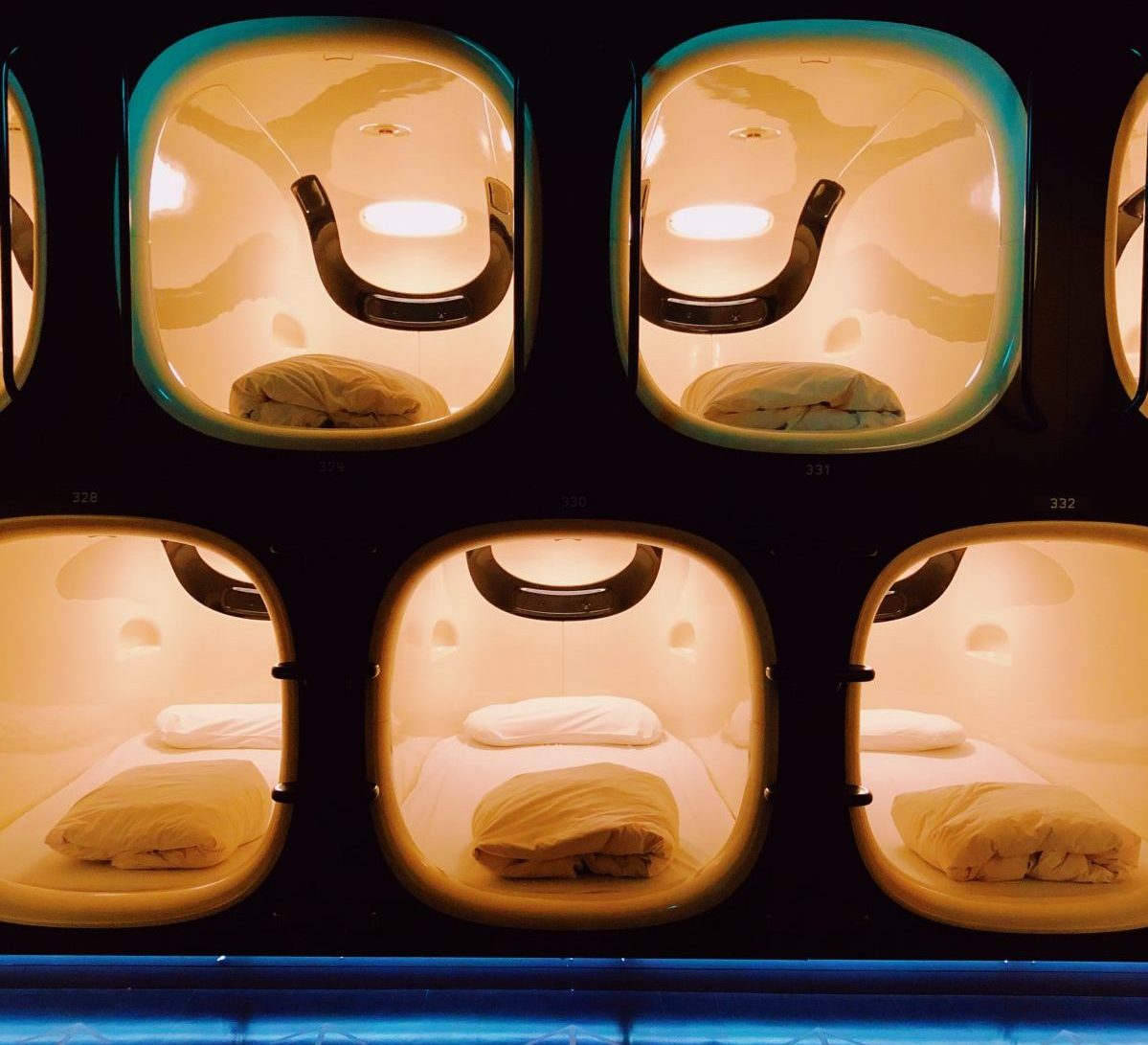 Photo of beds within small square-ish capsules with the blankets folded on top of the mattresses and the yellow lights on in each capsule, all built within a dark black wall and with capsule numbering on the floor