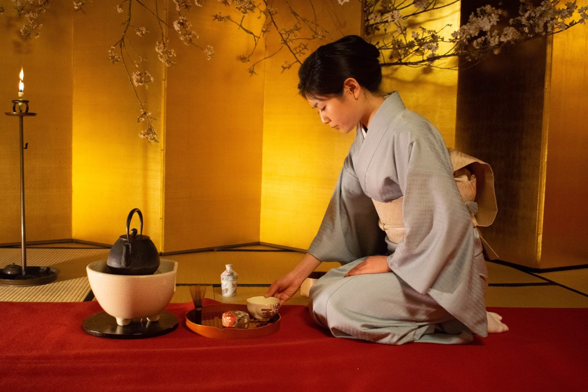 Photo of a woman sitting on her legs in a grey kimono with a cream colored sash grabbing a teacup from a circular platter and a black stone tea kettle on top of a bowl-shaped small table on a round black disc, all of these are on a red carpet with a golden ridged wall in the background, tree branches with small white flowers and a candle to the left side on a tall candle holder