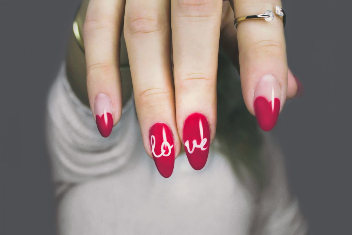 Photo of a woman’s fingers from the top with a ring on the index finger and the nails painted magenta, fully magenta with the word ‘love’ across the ring and middle finger, and heart-shaped magenta colors that fill half of the fingernail on the pinky and index fingers