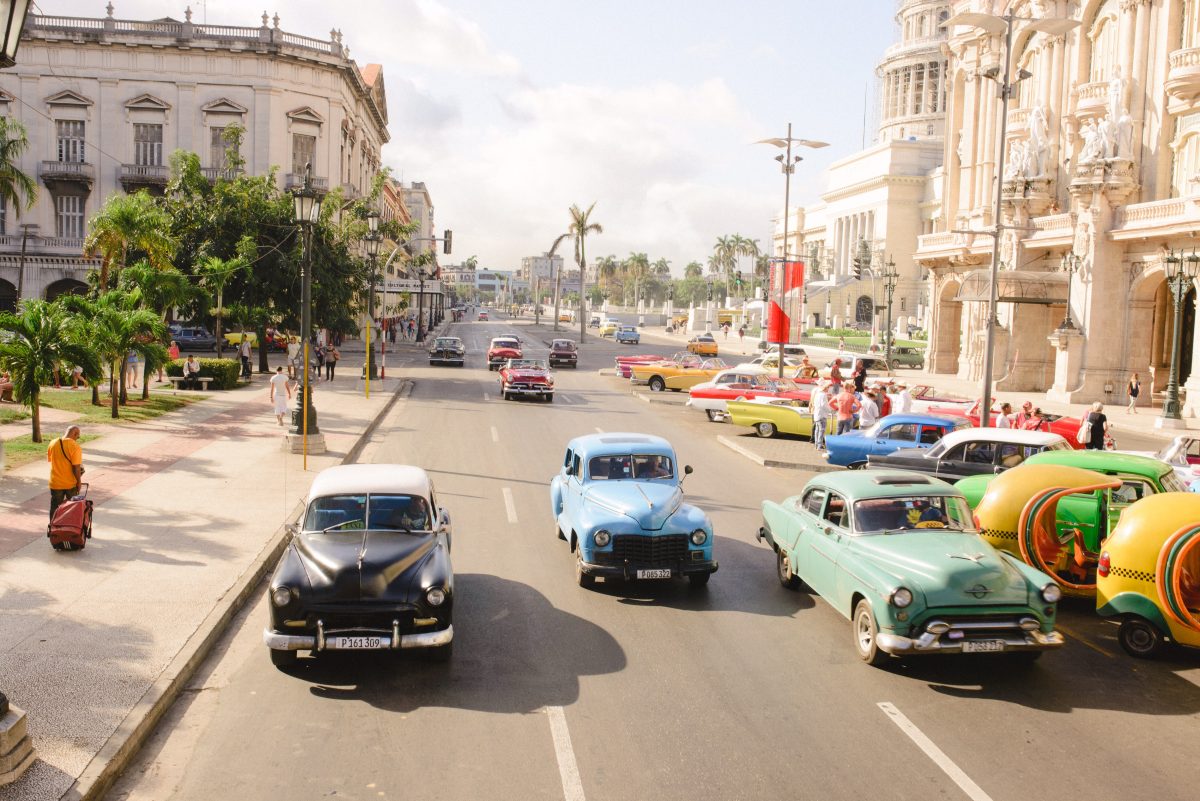 Photo of a wide road in Cuba with numerous colorful cars on the road that look retro with cream-colored Spanish style buildings on the blocks of the street with the city’s buildings and palm trees in the background with an afternoon cloudy sky