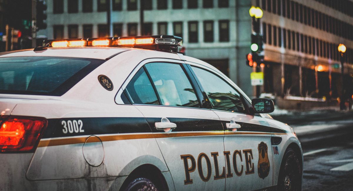 Photo of a dirty white police car with the word “POLICE” on the doors and its red sirens lit up with a blurred background of a big building and a city street