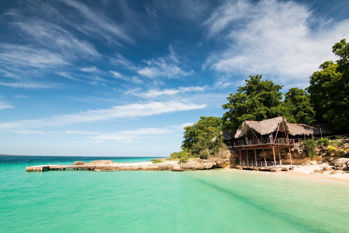 Photo of a turquoise-green Cuban beach with a ledge atop the water and a viewing beachfront local style structure And trees in the background with a cloudy sky