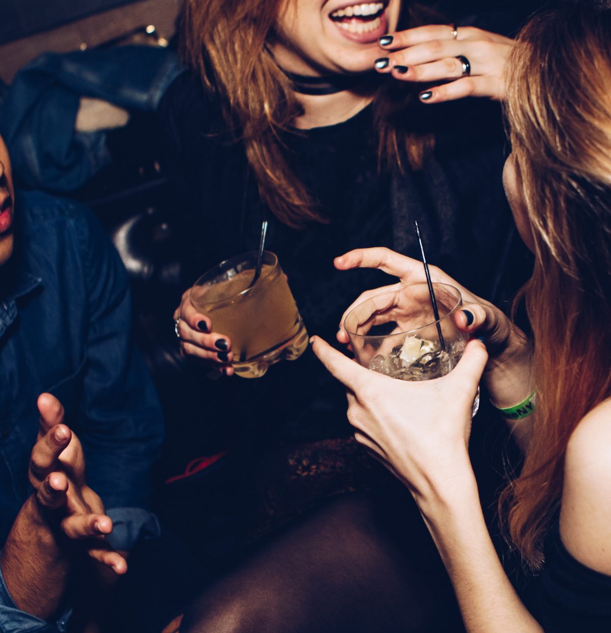 Photo of two women at a party talking while holding glasses with drinks and straws in them, both of them with brown mid-length hair, one woman’s back and arm is partially shown talking to the woman whose torso and lower half of the face also shown while smiling, and a guy next to the second woman barely seen in the shot