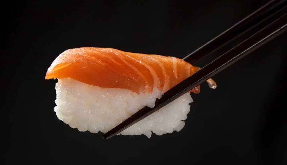 Salmon sushi being held by black chopsticks with a black background