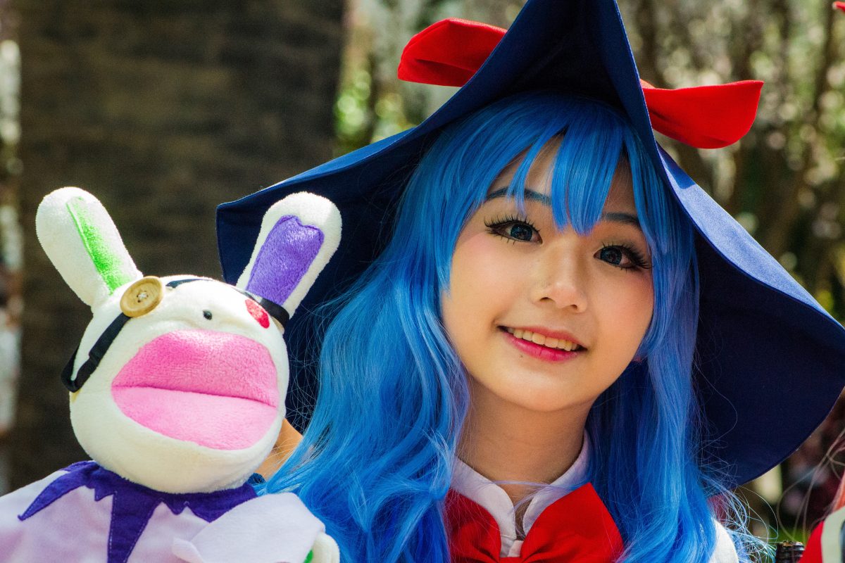 Photo of a girl with blue hair and makeup wearing a cosplay outfit with a big red ribbon below her neck and a big blue hat with a red ribbon while holding a white bunny with yellow and red buttons for eyes and big pink lips Photo of a girl with blue hair and makeup wearing a cosplay outfit with a big red ribbon below her neck and a big blue hat with a red ribbon while holding a white bunny with yellow and red buttons for eyes and big pink lips