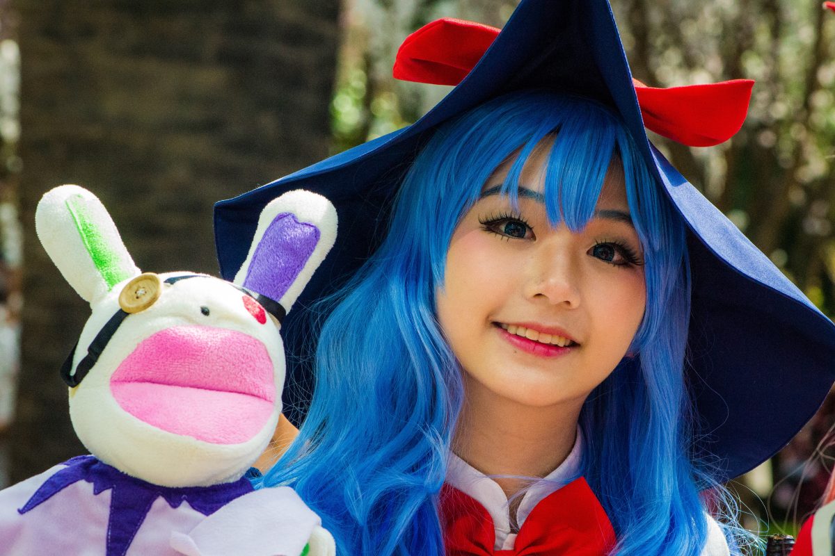 Photo of a girl with blue hair and makeup wearing a cosplay outfit with a big red ribbon below her neck and a big blue hat with a red ribbon while holding a white bunny with yellow and red buttons for eyes and big pink lips