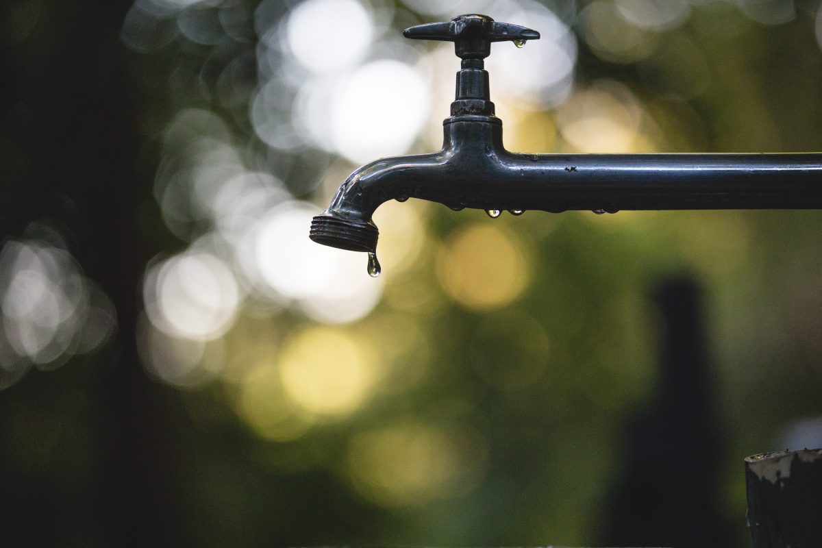 Photo of a metal tap with water dripping from it and the background is green and white that is blurred with a bokeh effect