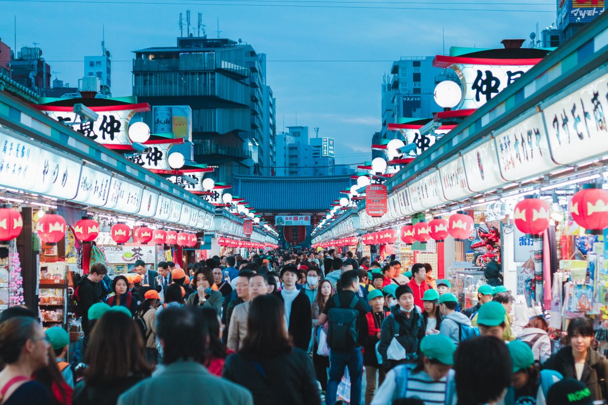 Photo of a busy night market with both left and right side lined with stalls next to each other with indistinct sign designs and red lanterns hanging from each stall, with Japanese style lights on top of the stalls and a busy crowd walking along the entire night market, with a view of buildings and a traditional style small Japanese structure at the end in the background