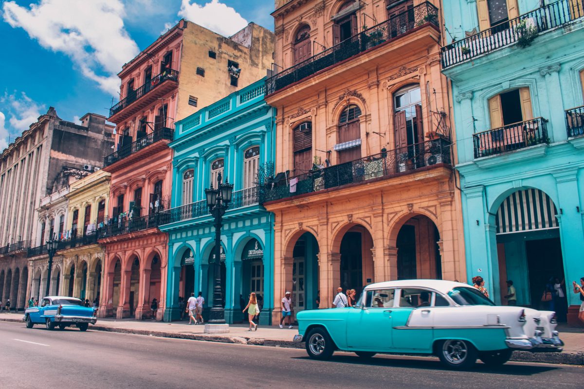 Photo of colorful Cuban buildings with orange, blue, and cream with archways in front of the buildings’ entryways and balconies and blue retro style cars along the streets and people walking on the sidewalk, with the weather being bright and cloudy