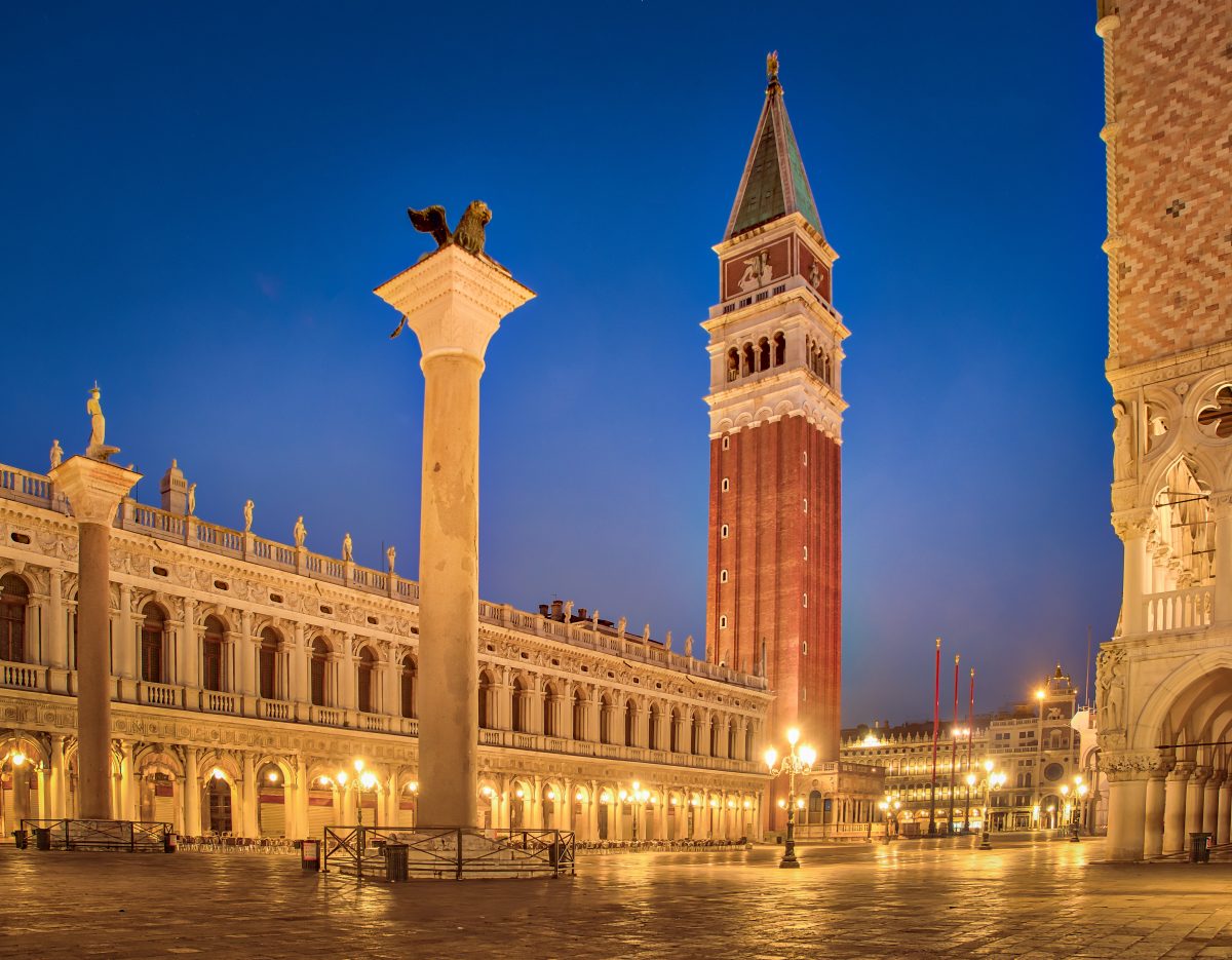 Photo of San Marco at night with a wide area with no people and Venetian buildings surrounding it with a tall tower at the center of the plaza with an animal statue on top, and in the near distance a tall building with a pointed tip is seen, with street lamps giving the area bright light Photo of San Marco at night with a wide area with no people and Venetian buildings surrounding it with a tall tower at the center of the plaza with an animal statue on top, and in the near distance a tall building with a pointed tip is seen, with street lamps giving the area bright light