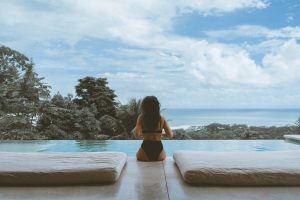Photo of the backside of a woman with long hair wearing a black two-piece swimsuit sitting on the edge of an infinity pool with two mattresses on either side of her, beyond the pool are the tops of trees and a distant view of the ocean with a cloudy blue sky