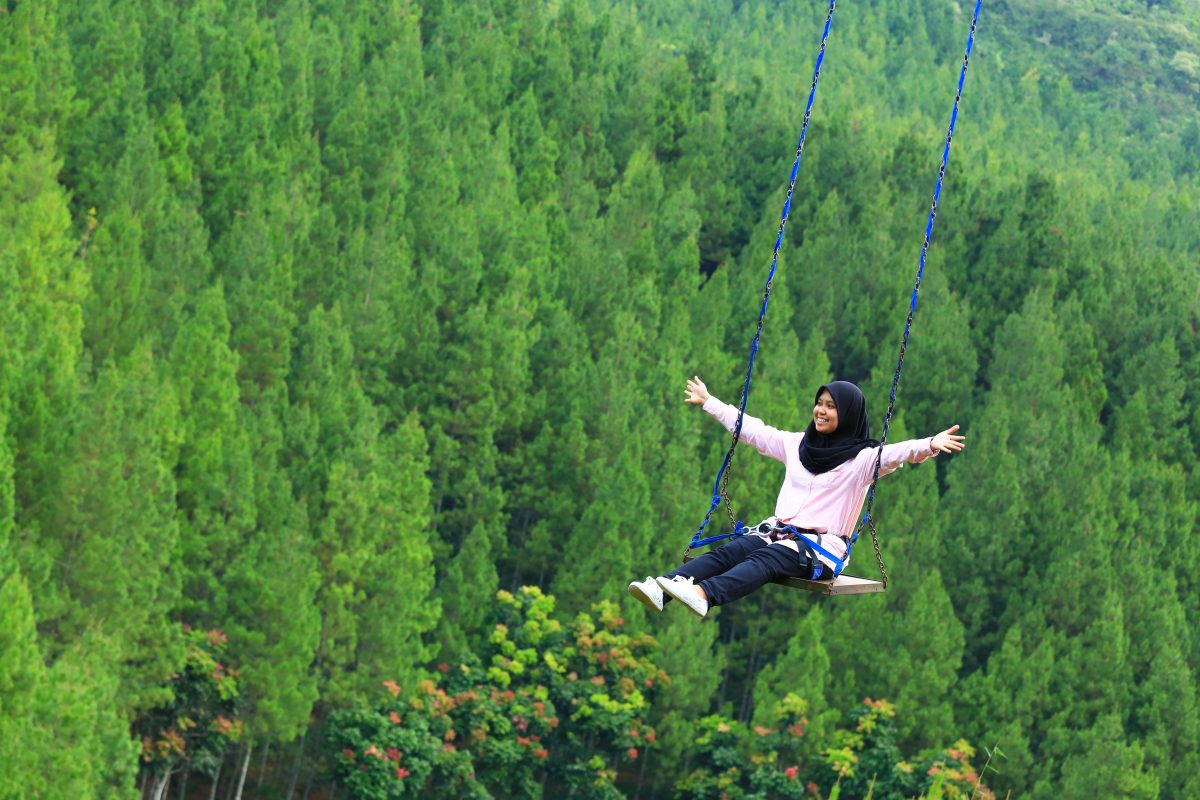 A woman swings over trees with open arms and a wide smile in Maribaya, Bandung, Indonesia