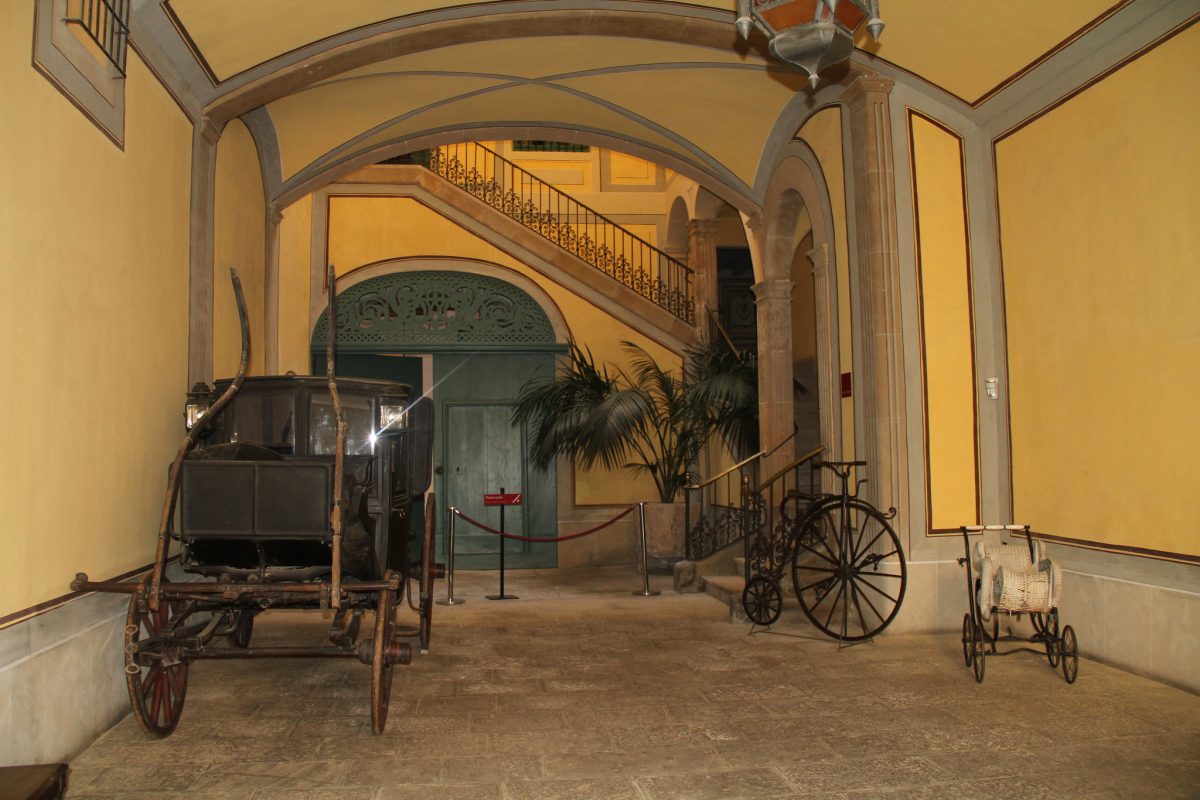 An old carriage, bike, and stroller are displayed in front of a staircase at Museu Romantic in Sitges, Spain