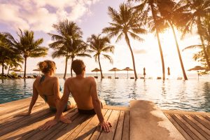 The perfect Hawaii honeymoon, a couple lounging by the pool overlooking the ocean and coconut palm trees at sunset.