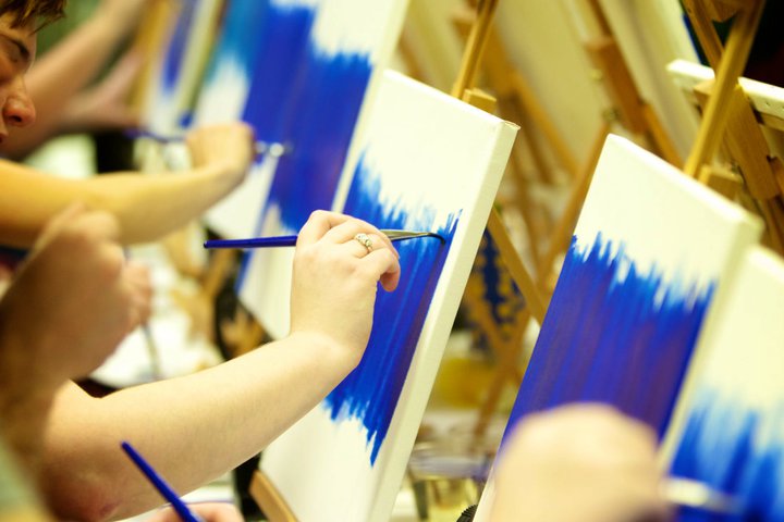 Close-up of hands holding paint brushes against a white canvas with blue paint. Close-up of hands holding paint brushes against a white canvas with blue paint.