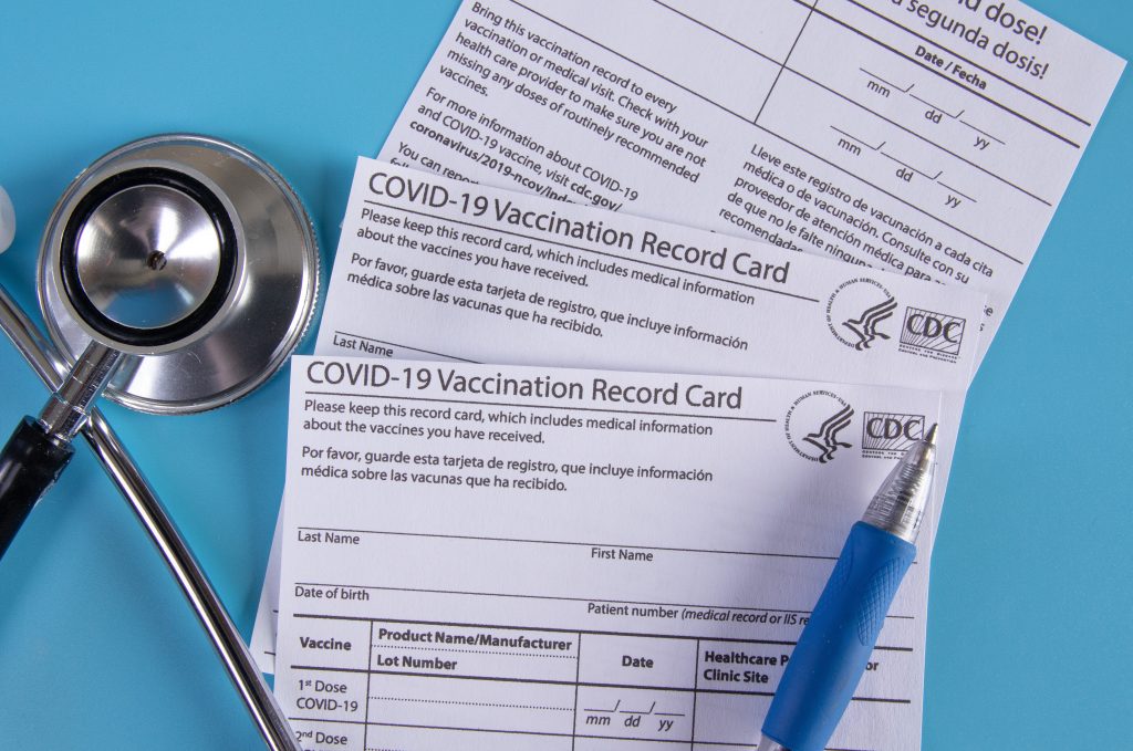 Three blank COVID-19 vaccination record cards with a stethoscope and pen on a blue background.