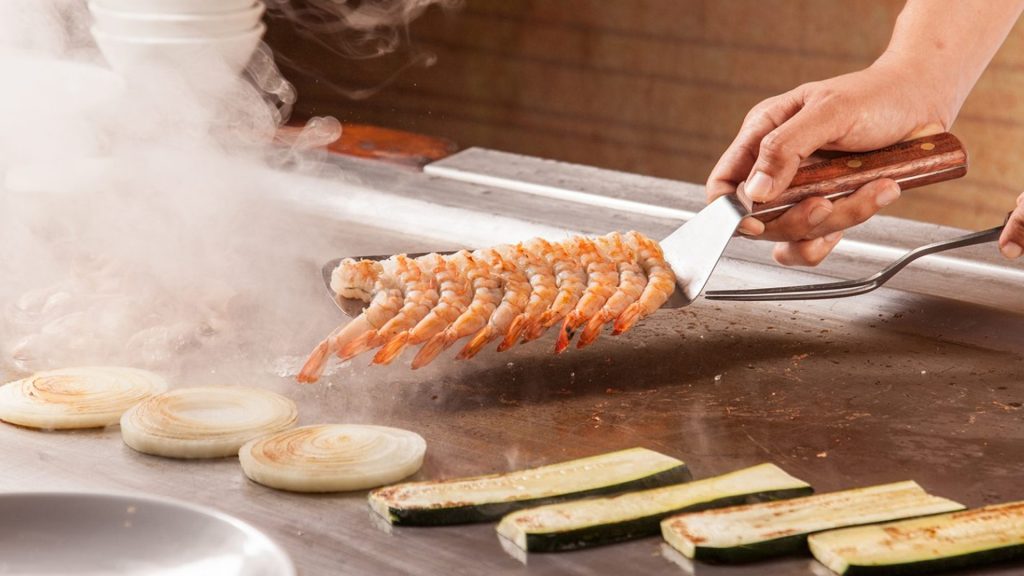 Close-up shot of a hand grilling shrimp, onions, and eggplants on a hibachi grill.