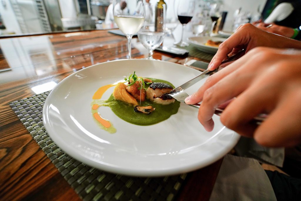 Close-up of hands using a knife and fork to eat scallops with a spinach puree. 