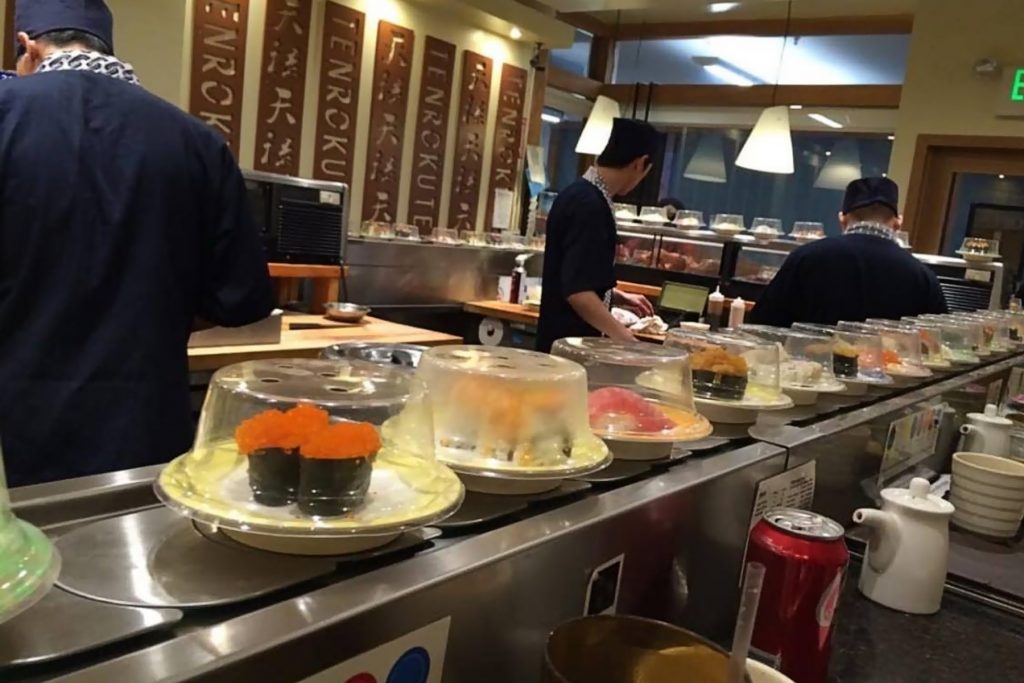 Plates of sushi on a conveyor belt with three chefs in the background preparing food at Tenroku Sushi.