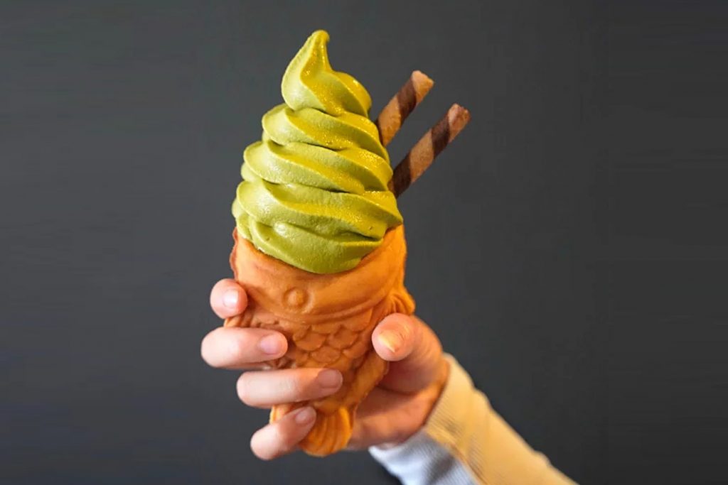Close-up shot of a hand holding a matcha soft-serve ice cream on a fish-shaped cone with wafers sticks.