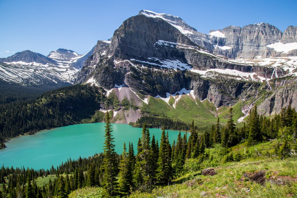 Wide shot of a lake and greenlands at the Glacier National Park with snow-capped mountain range in the background. 