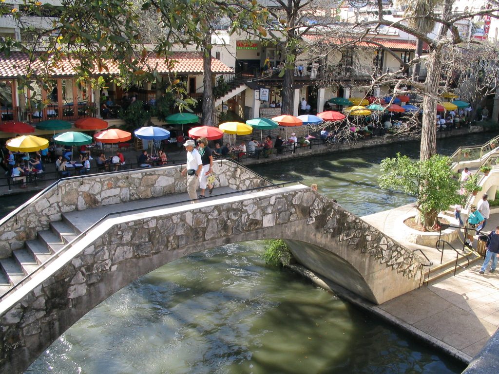 Couple walking across the bridge at the San Antonio RiverWalk in Texas, with colorful umbrellas lined in the background.