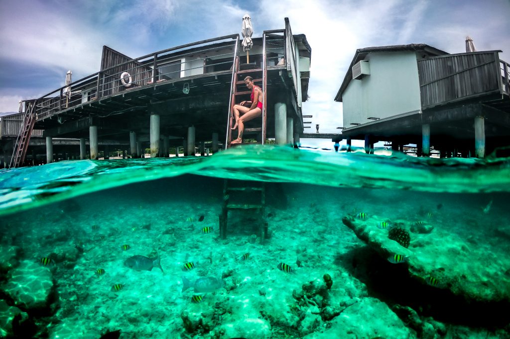 Split view of a woman sitting on a ladder near the edge of water in one of the bungalows in Maldives, with fish swimming below the water.