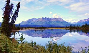 Reflection of a mountain range, surrounding greenery, and the sky in a body of water in Alaska.