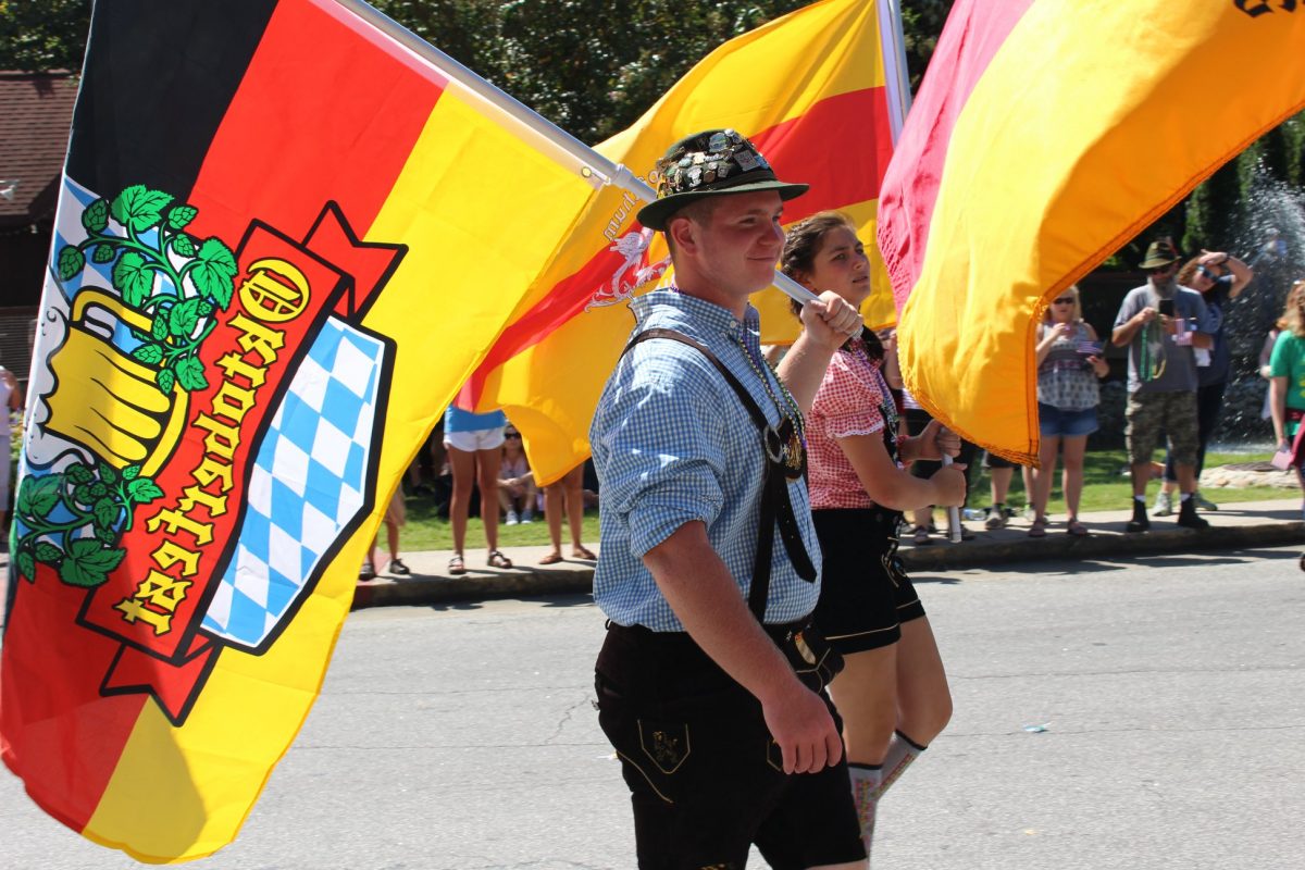 Man wearing lederhosen holding an Oktoberfest flag during the Helen Oktoberfest parade.