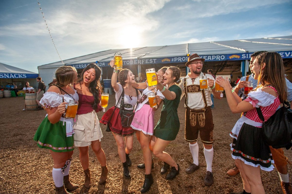 Group wearing dirndls and lederhosen dancing while holding beer mugs during Oktoberfest in California.