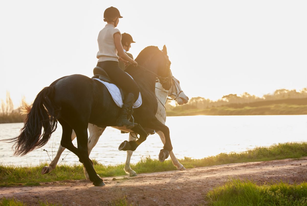 Two women on horseback on a trail, near a body of water.