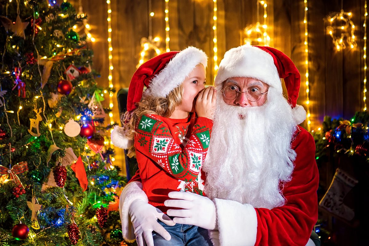 Little girl whispering to Santa while sitting on his lap.