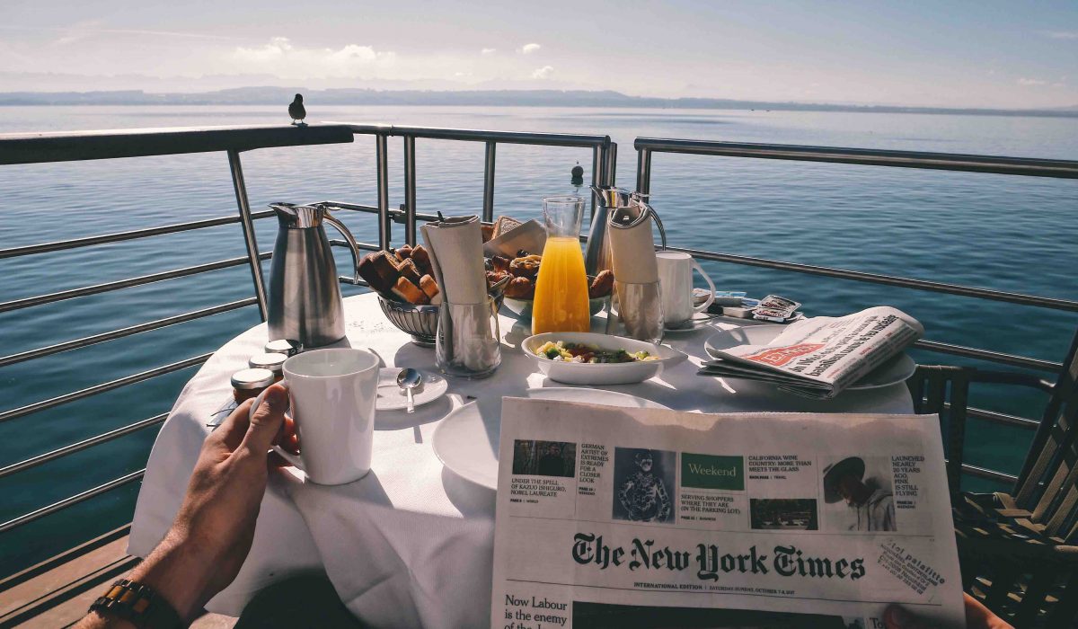 cruise deck reading a newspaper at a breakfast table with various food and beverages on a deck with a seaview