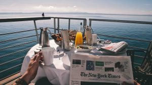 reading a newspaper at a breakfast table with various food and beverages on a deck with a seaview