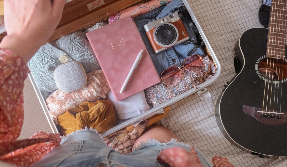 a top view shot of a luggage filled with clothes, a pink diary, a camera and a pair of shades