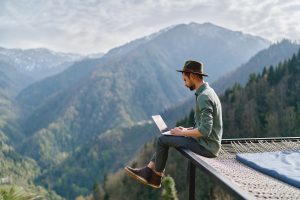 Young man freelancer traveler wearing hat anywhere working online using laptop and enjoying mountains view
