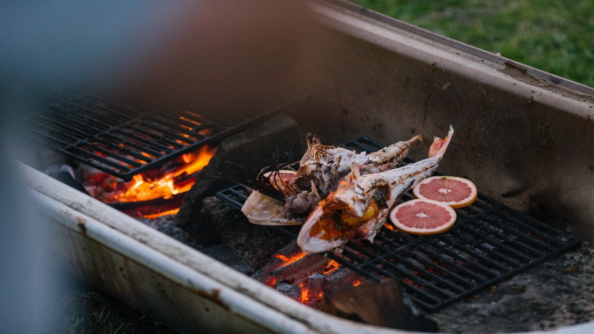 Two open-bellied fish being grilled with cut citrus fruit also grilling next to the fish and the grill on top of a fire lit by coal within a grilling basin