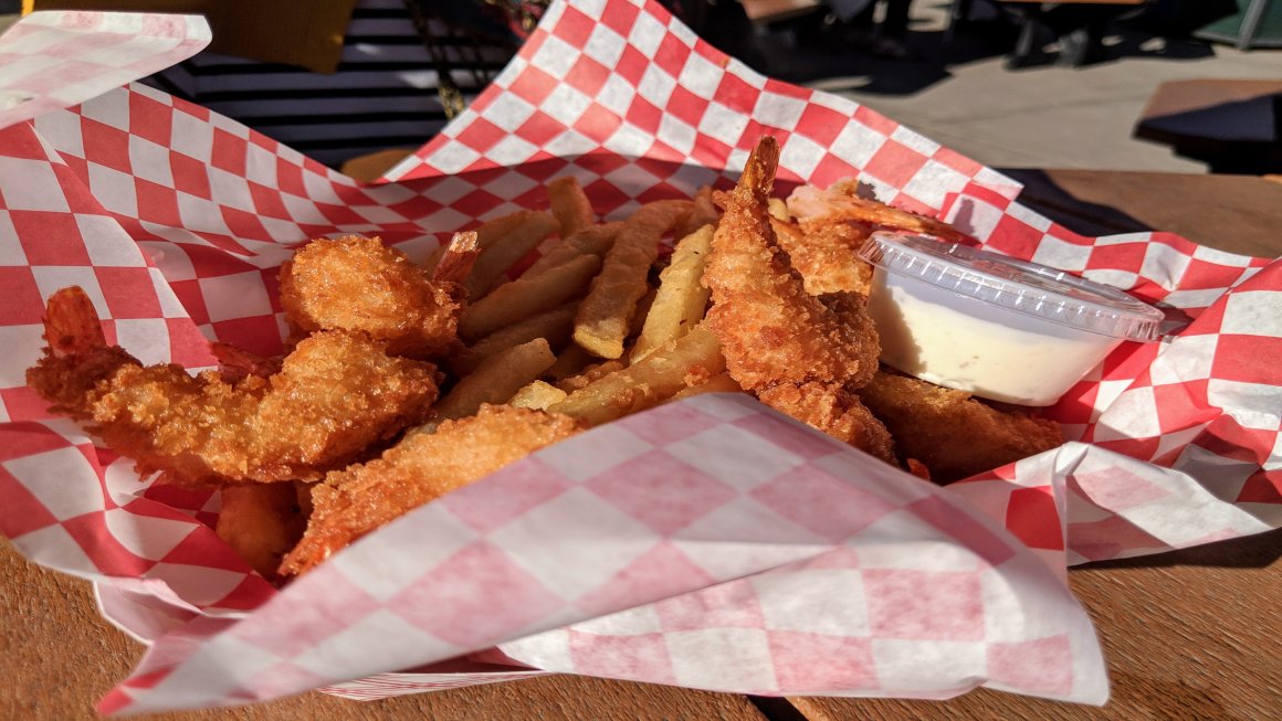 Deep-fried fish and chips with a sauce in a container and all of them contained within red and white checkered paper on a wooden brown table