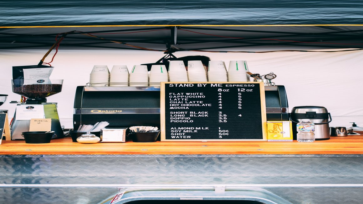 Photo of a Salamanca Market Coffee stand with cups and the coffee maker and the menu all in sight on a wooden and metal table with a white backdrop and a dark blue cover on top