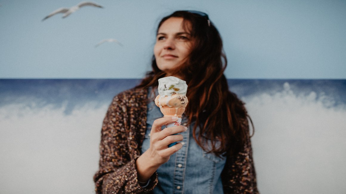 Photo of a woman with long brown hair wearing a denim polo and a brown patterned cardigan holding an ice cream cone with white and peach scoops melting with a painted blue background of a sky with birds and a blue ocean