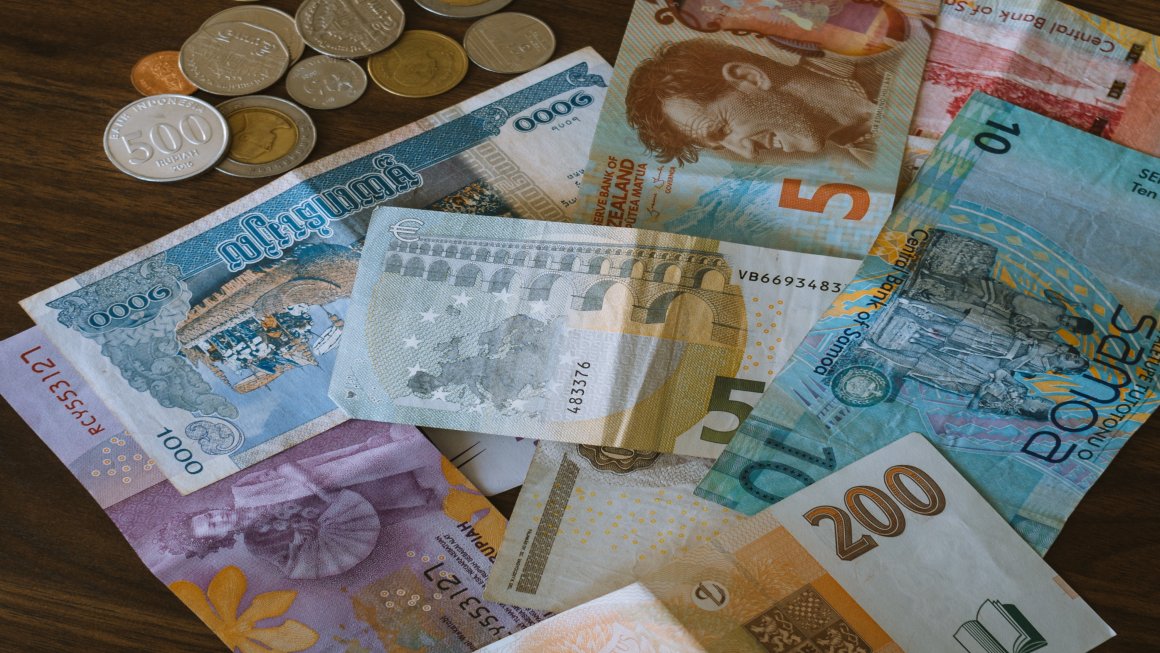 Photo of different paper money currencies on a brown wooden table, from 5s to 10s, to 200s and so on, with many different colors and designs and coins in the upper left corner of the shot
